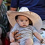 baby, child, cowboy_hat, muddy_hat, onesie, person, hand, adult_hand, face, curious, seated, indoor, casual_clothing, people, portrait, blurry_background, holding, light_skin, short_hair, expression