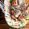 baby, baby_rocker, child, indoor, harness, striped_clothing, colorful_pattern, wooden_floor, rug, infant, seat, person, cute, comfort, looking_at_camera, small_hands, small_feet, calm_expression, soft_lighting, home