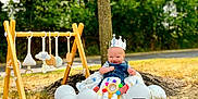 Easton joined the competition — help win amazing prizes! baby, balloons, blanket, celebration, child, clothing, crown, grass, happy, headwear, nature, outdoor, park, person, play_gym, smiling, sunlight, toy, tree, wooden_frame