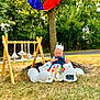 baby, crown, balloons, blanket, outdoor, grass, tree, toy, play_gym, smiling, child, sunlight, wooden_frame, nature, celebration, happy, clothing, person, headwear, park