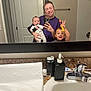 woman, baby, toddler, bathroom, mirror, selfie, toothbrush, sink, countertop, soap_dispenser, door, towel_rod, smile, person, reflection, casual, clothing, playful, indoor, family