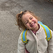 Ghita a rejoint le concours — aidez-le/la à gagner de superbes lots ! child, smiling, girl, curly_hair, backpack, coat, pink_boots, concrete, pavement, outdoor, joyful, portrait, candid, happy, teeth, playful, urban, shrubbery, winter_clothing, school_age