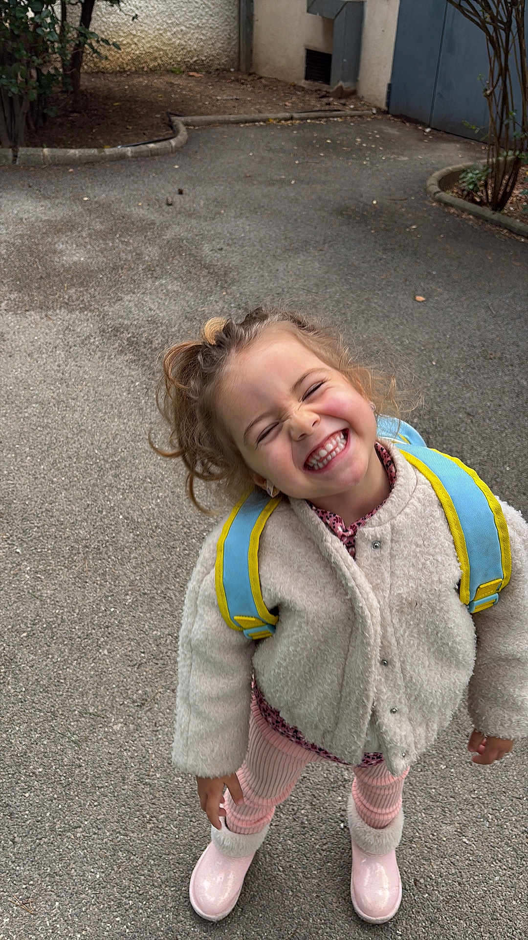 Ghita a rejoint le concours — aidez-le/la à gagner de superbes lots ! child, smiling, girl, curly_hair, backpack, coat, pink_boots, concrete, pavement, outdoor, joyful, portrait, candid, happy, teeth, playful, urban, shrubbery, winter_clothing, school_age