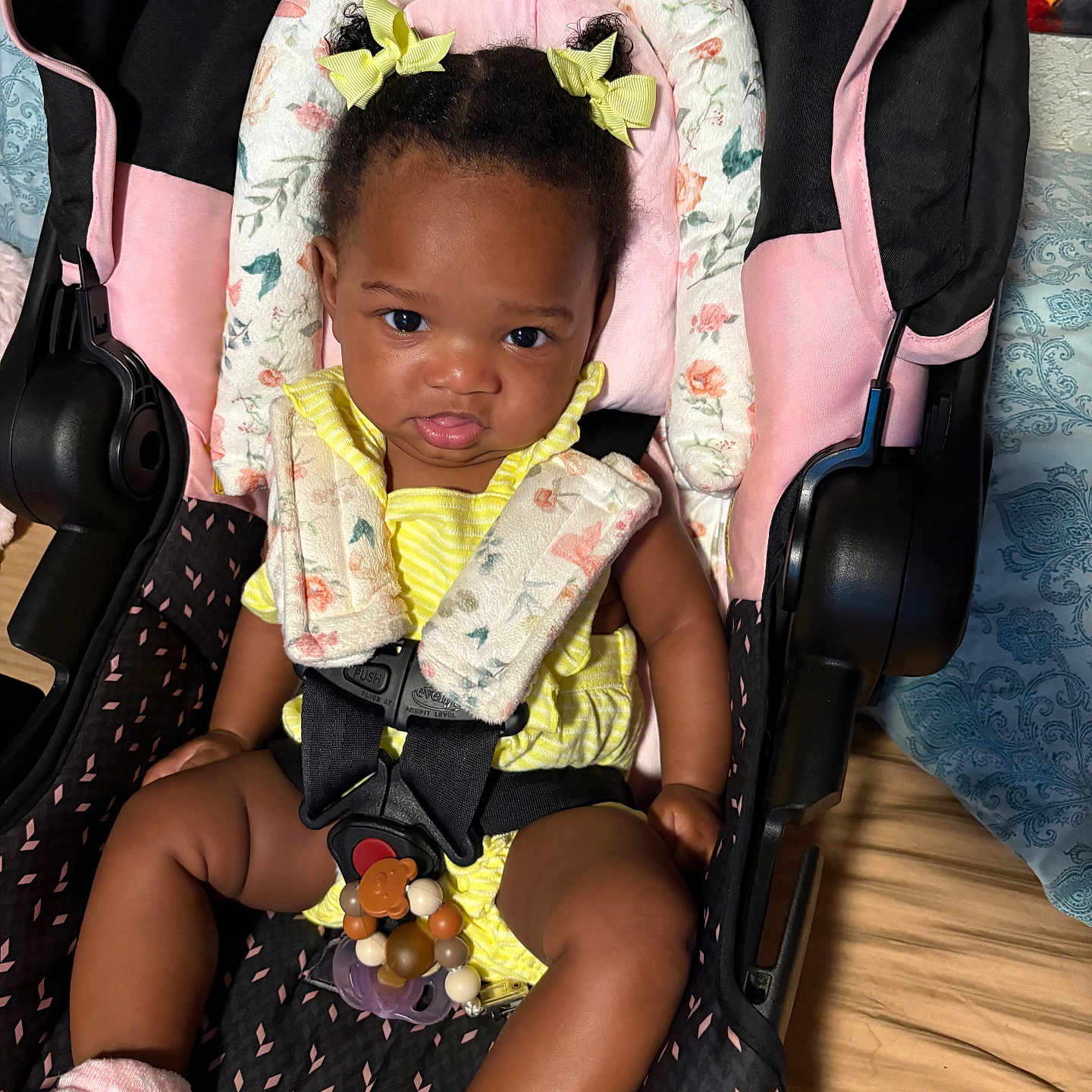 Majesti is registered to the contest to win money with this photo: baby, black_hair, blanket, car_seat, child, clothing, curious_expression, cute, floral_pattern, indoor, infant, person, pink_socks, portrait, seatbelt, straps, toddler, wooden_floor, yellow_bows, yellow_outfit