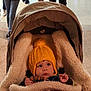 baby, child, stroller, blanket, hat, yellow_hat, pom_pom, toy, wooden_beads, face, eyes, hand, clothing, warm, indoor, floor, people, walking, person, curious