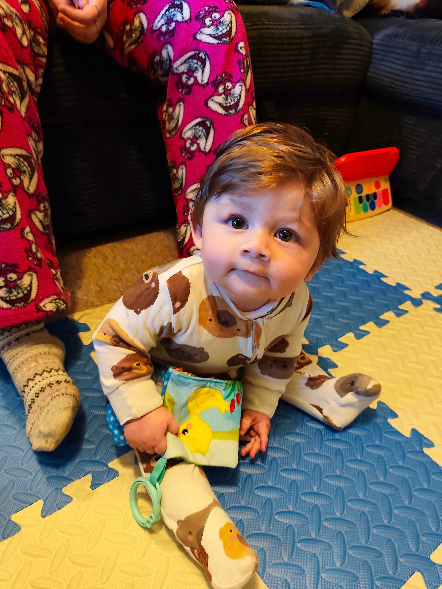 Eric is registered to the contest to win money with this photo: baby, child, onesie, toy, floor_mat, pajamas, socks, person, indoor, carpet, couch, hand, face, smile, cute, play, colorful, soft_toy, floor, seated