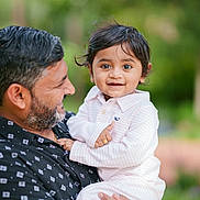 Jihan Patel joined the competition — help win amazing prizes! child, toddler, man, adult, holding, smiling, outdoor, portrait, person, black_hair, beard, casual_clothing, striped_shirt, ring, happy, nature, blurred_background, family, affection, closeup
