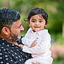 child, toddler, man, adult, holding, smiling, outdoor, portrait, person, black_hair, beard, casual_clothing, striped_shirt, ring, happy, nature, blurred_background, family, affection, closeup