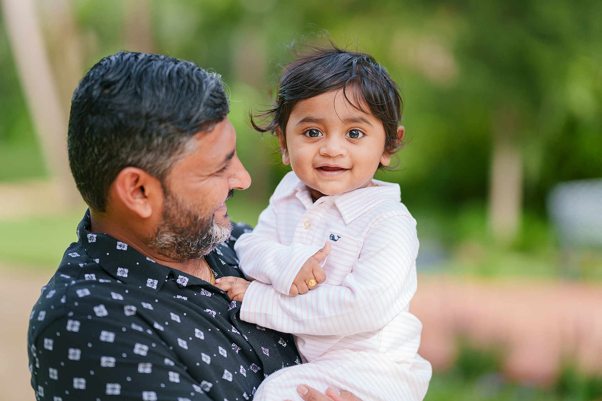 Jihan Patel joined the competition — help win amazing prizes! child, toddler, man, adult, holding, smiling, outdoor, portrait, person, black_hair, beard, casual_clothing, striped_shirt, ring, happy, nature, blurred_background, family, affection, closeup