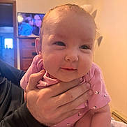 Amarianna joined the competition — help win amazing prizes! baby, infant, child, person, hand, pink_clothing, face, indoor, furniture, television, living_room, smile, cute, human, skin, arm, adult_hand, holding, soft_light, cozy