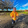 child, toddler, yellow_jacket, raincoat, outdoor, tree, bare_tree, blue_sky, grass, rural, standing, sneakers, shoes, pavement, shadow, sunlight, portrait, face, ground, playful_expression