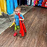 Kai is registered to the contest to win money with this photo: toddler, child, toy, baseball_bat, clothing, store, aisle, flooring, shoes, smiling, person, blue_shirt, yellow_shorts, red_shoes, hanging_clothes, retail, indoor, shopping, plastic_toy, happy