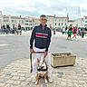man, dog, leash, muzzle, street, cobblestone, people, buildings, outdoor, cloudy_sky, casual_clothing, glasses, plant_pot, walking, urban, sidewalk, public_space, daytime, pedestrians, bicycle