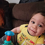 Ricardo is registered to the contest to win money with this photo: toddler, child, toy, yellow_shirt, striped_pants, couch, brown_furniture, indoor, smiling, face, eyes, playful, person, baby, clothing, home, relaxing, cute, infant, casual