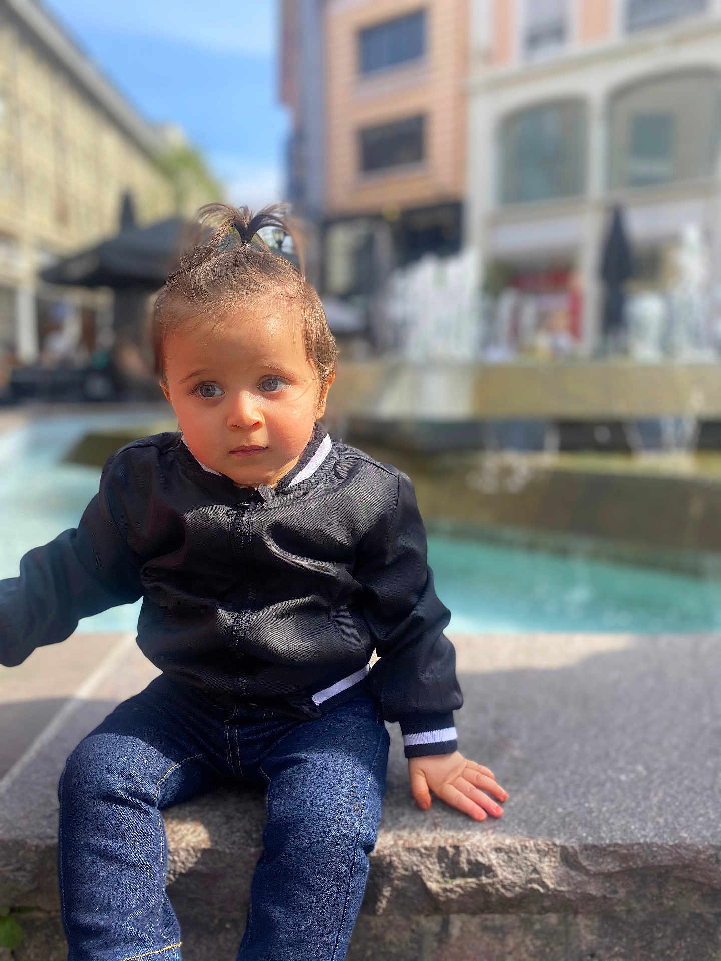 Ziyad participe au concours pour gagner de l'argent avec cette photo : toddler, child, baby, jeans, jacket, pony_tail, outdoor, fountain, stone_ledge, urban, cityscape, sunlight, person, casual_clothing, young_child, sitting, portrait, daytime, background_blur, cute