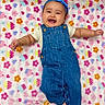 baby, smiling, denim_overalls, headband, blue, blanket, floral_pattern, happy, infant, cute, lying_down, joyful, small_arms, adorable, cheerful, child, soft_texture, colorful, playful, portrait