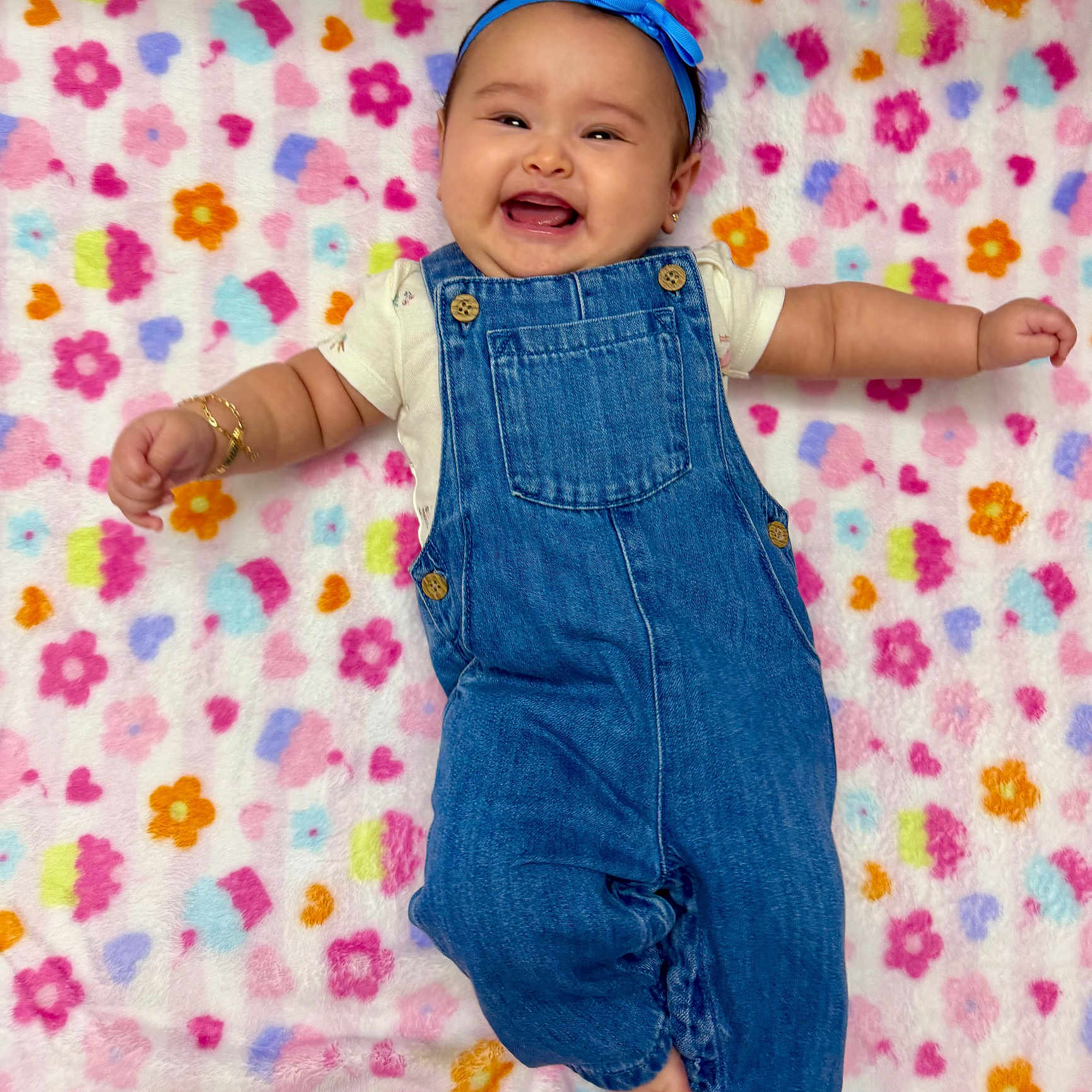 Valerie is registered to the contest to win money with this photo: baby, smiling, denim_overalls, headband, blue, blanket, floral_pattern, happy, infant, cute, lying_down, joyful, small_arms, adorable, cheerful, child, soft_texture, colorful, playful, portrait