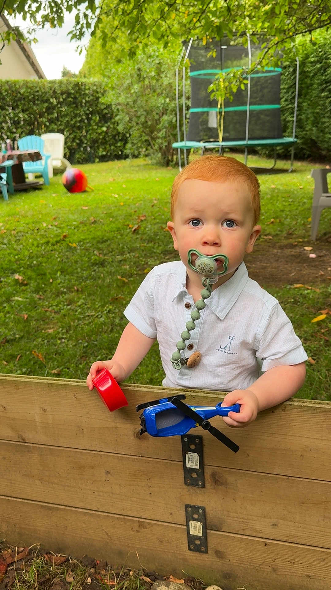 Augustin participe au concours pour gagner de l'argent avec cette photo : toddler, child, pacifier, toy_helicopter, wooden_fence, grass, garden, outdoor, greenery, chair, table, trampoline, red_hair, white_shirt, play, curious, young_child, daylight, backyard, nature