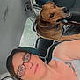 woman, glasses, tattoo, dog, car_interior, seat, headrest, window, seatbelt, smile, pet, brown_dog, casual_clothing, portrait, daylight, vehicle, backseat, human, companion, road