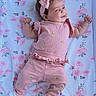baby, infant, pink_outfit, headband, bow, smile, lying_down, blanket, floral_pattern, cute, child, soft_lighting, indoors, happy, adorable, person, small_hands, cozy, playful, toddler