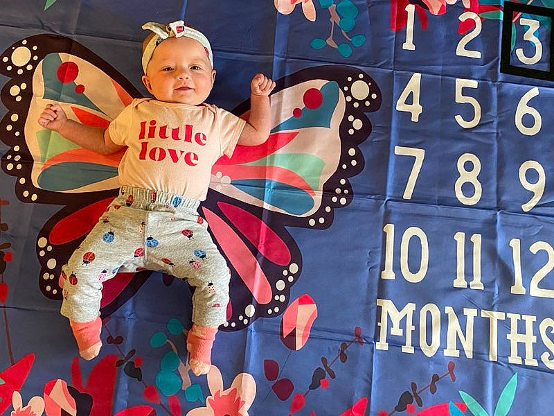 Izzy is registered to the contest to win money with this photo: advertising, baby_toddler_clothing, carmine, event, fictional_character, font, fun, games, graphics, happy, logo, number, pattern, person, recreation, rectangle, sitting, t_shirt, textile, toddler