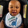 baby, child, smiling, car_seat, bib, dinosaur, happy, infant, clothing, black_shirt, face, portrait, cute, hands, closeup, expression, seatbelt, warning_label, indoors, person