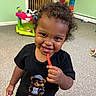 Casimir is registered to the contest to win money with this photo: child, toddler, smiling, happy, curly_hair, portrait, snack, red_candy, holding_food, black_shirt, graphic_tshirt, teddy_bear, playroom, toys, colorful, indoor, carpet, crib, storage_pockets, closeup