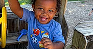 Casimir joined the competition — help win amazing prizes! child, toddler, smiling, happy, outdoor, playground, wooden_playhouse, play_structure, raised_hand, curly_hair, blue_shirt, gray_pants, shoes, toy_shovel, trees, grass, sunlight, portrait, summer, playtime