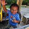 child, toddler, smiling, happy, outdoor, playground, wooden_playhouse, play_structure, raised_hand, curly_hair, blue_shirt, gray_pants, shoes, toy_shovel, trees, grass, sunlight, portrait, summer, playtime