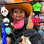 baby, cowboy_hat, shopping_cart, plush_toy, mickey_mouse, tattoo, bib, smiling, child, store, halloween_mask, costume, colorful_hats, black_clothing, indoor, happy, toy, person, retail, cute