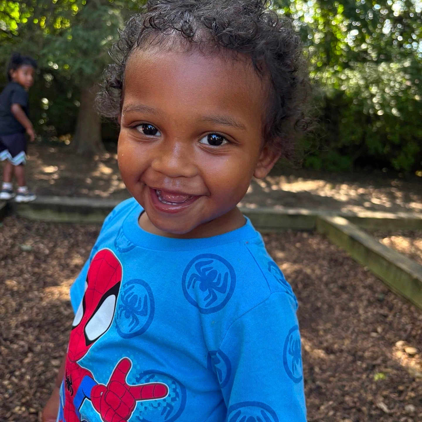 Casimir is registered to the contest to win money with this photo: child, toddler, smiling, happy, outdoor, playground, portrait, spiderman, superhero, blue_shirt, curly_hair, dark_skin, close_up, standing, trees, nature, bokeh, background_child, play_area, daylight