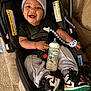 baby, smiling, car_seat, grey_hat, sweatpants, sneakers, bottle, pacifier_clip, happy, indoor, child, footwear, toy, seatbelt, person, cute, infant, clothing, accessory, cozy