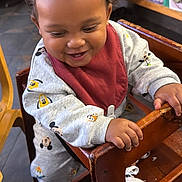 Casimir is registered to the contest to win money with this photo: baby, bib, casual, chair, child, clothing, curly_hair, cute, happy, high_chair, indoor, mickey_mouse, person, playful, restaurant, shoes, smiling, table, toddler, wooden_furniture