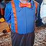 child, costume, orange_hat, orange_glasses, orange_bow_tie, orange_suspender, blue_shirt, gray_pants, barefoot, smiling, indoor, carpet, toy, water_bottle, person, pants, feet, happy, glasses, floor