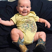 Urijah is registered to the contest to win money with this photo: baby, child, smile, onesie, yellow_clothing, cowboy_boots, footwear, sitting, indoor, quilted_blanket, happy, cute, portrait, infant, person, face, skin, toy_story, clothing, expression