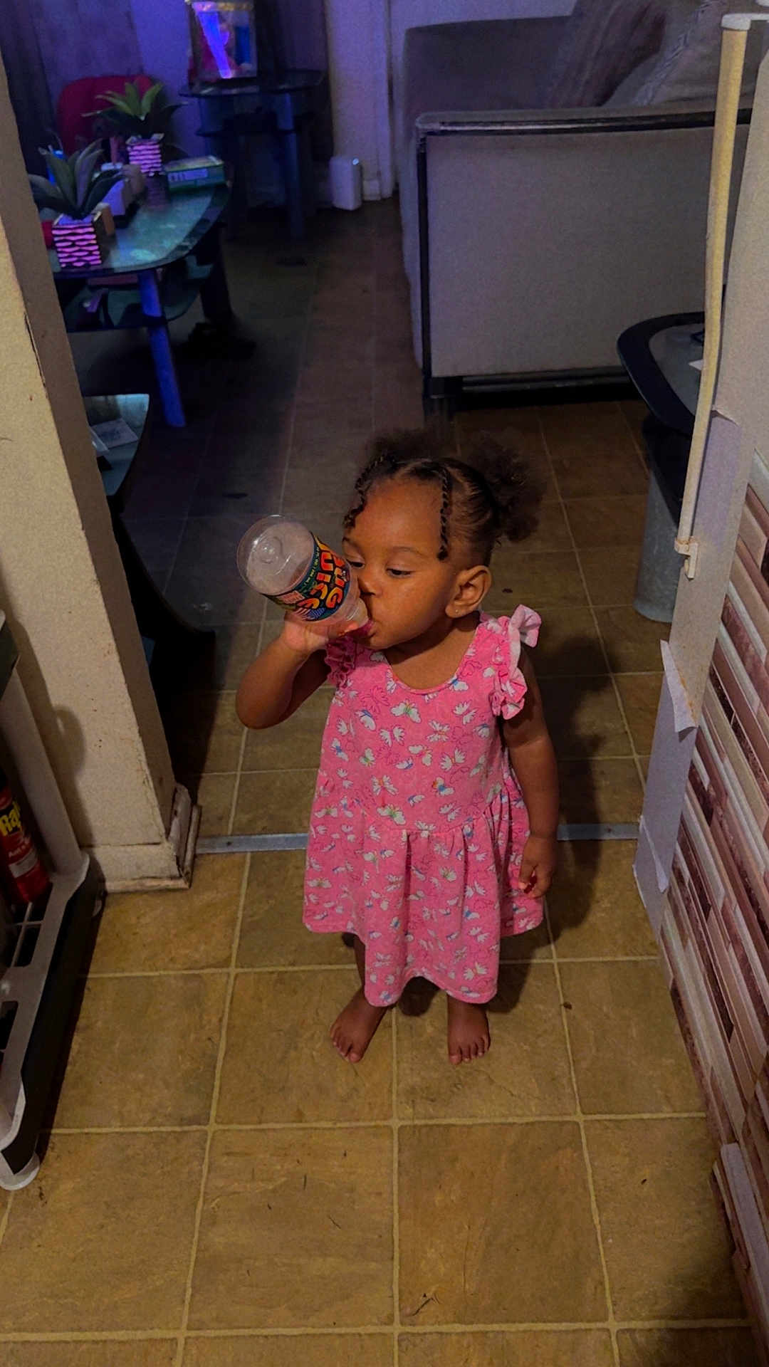 Hazel joined the competition — help win amazing prizes! toddler, child, pink_dress, barefoot, indoor, drinking, bottle, floor, tile, hair_puffs, furniture, couch, table, plant, book, person, home, concentration, young_child, casual