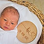 newborn, baby, infant, blanket, blue_ribbon, basket, woven_basket, wooden_plaque, text, face, head, eyes, skin, swaddled, white_blanket, portrait, indoors, cute, announcement, calm