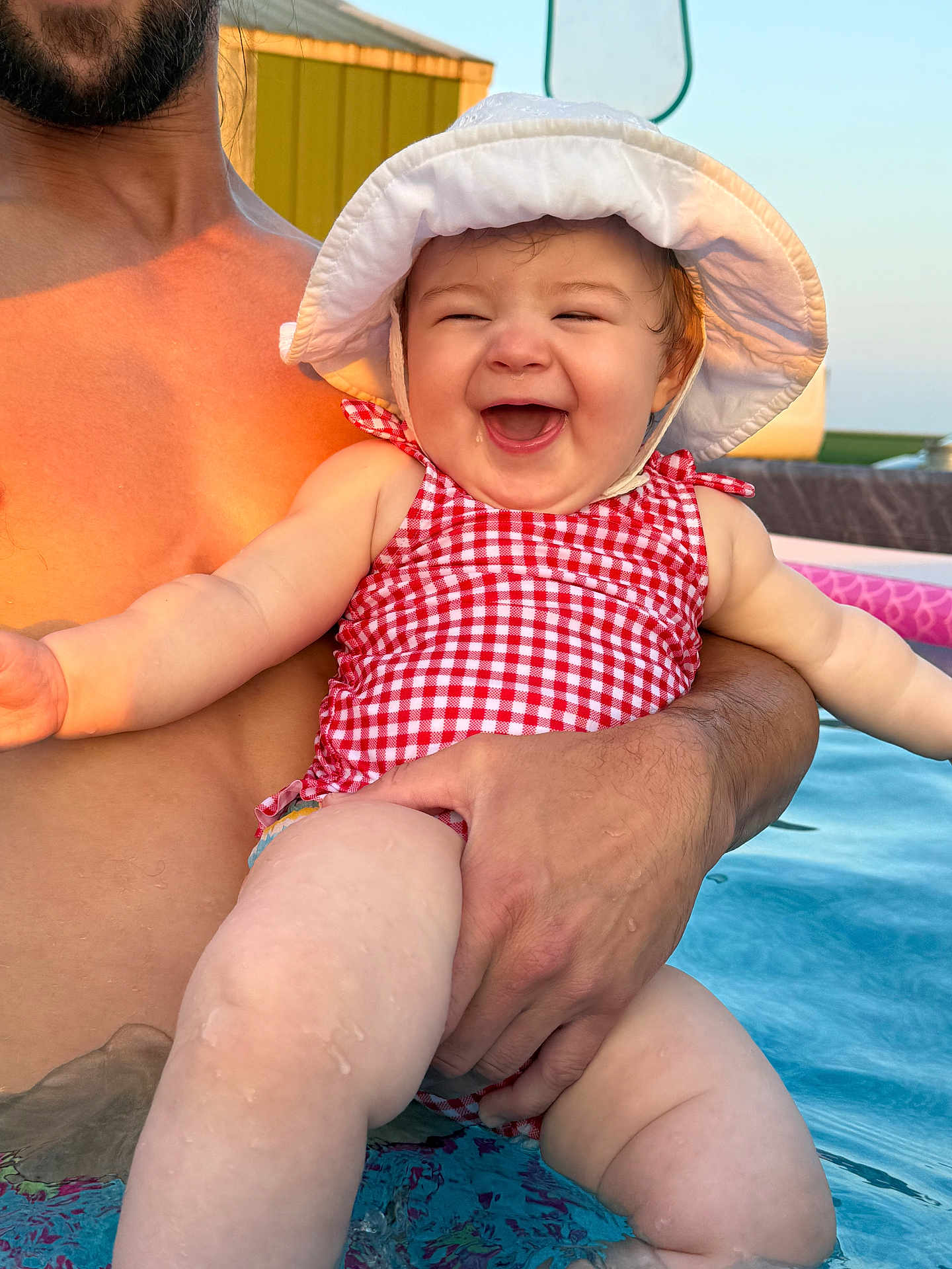 Stevie is registered to the contest to win money with this photo: adult_hand, arm, baby, checkered_pattern, child, fun, happy, laughing, leg, outdoor, pool, skin, smiling, summer, sun_hat, sunlight, swimsuit, vacation, warm_light, water