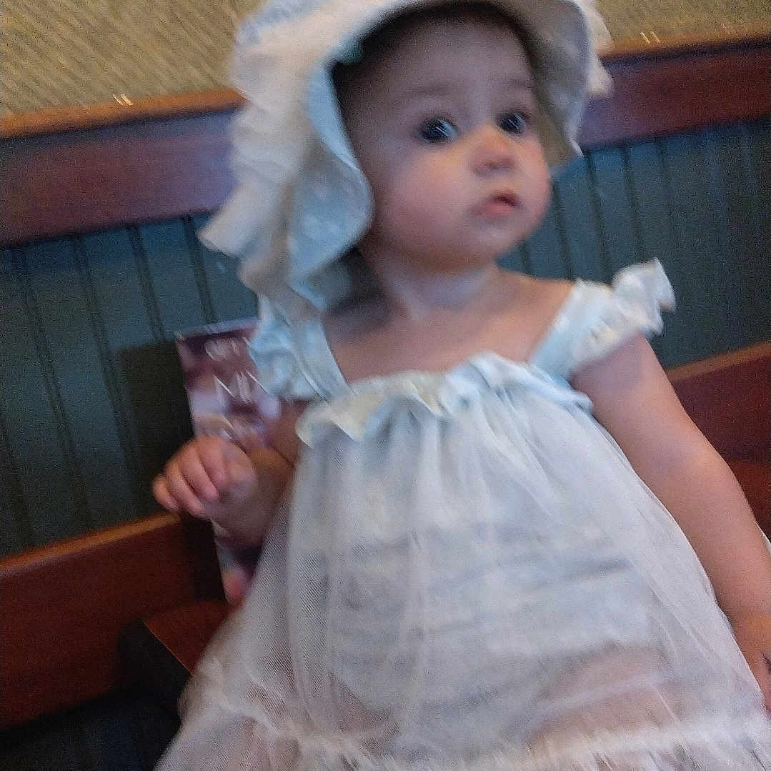 Kamryn is registered to the contest to win money with this photo: baby, bodypart, bonnet, clothing, costume, dress, eveningdress, face, finger, furniture, hand, hardwood, hat, head, person, photography, portrait, sitting, sunhat, wood