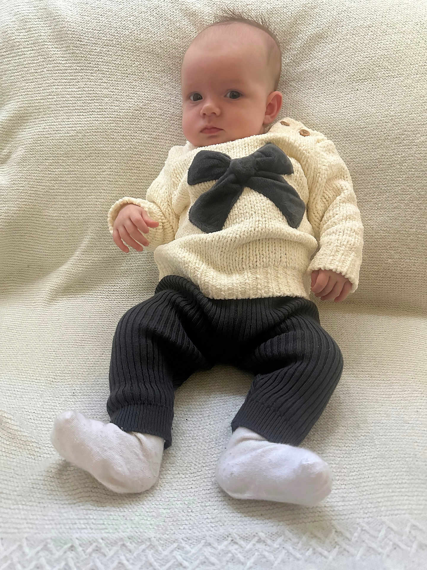 Lottie is registered to the contest to win money with this photo: baby, infant, child, sweater, bow, black_bow, pants, white_socks, knitted_clothing, textured_blanket, seated, indoors, portrait, cute, young_child, looking_at_camera, cozy, comfortable, soft_lighting, neutral_colors