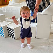 Beau is registered to the contest to win money with this photo: toddler, child, baby, indoor, play_area, white_socks, bow_tie, suspenders, smiling, holding_hand, adult, soft_blocks, checkered_pattern, floor_mat, shorts, cute, walking, person, happy, light