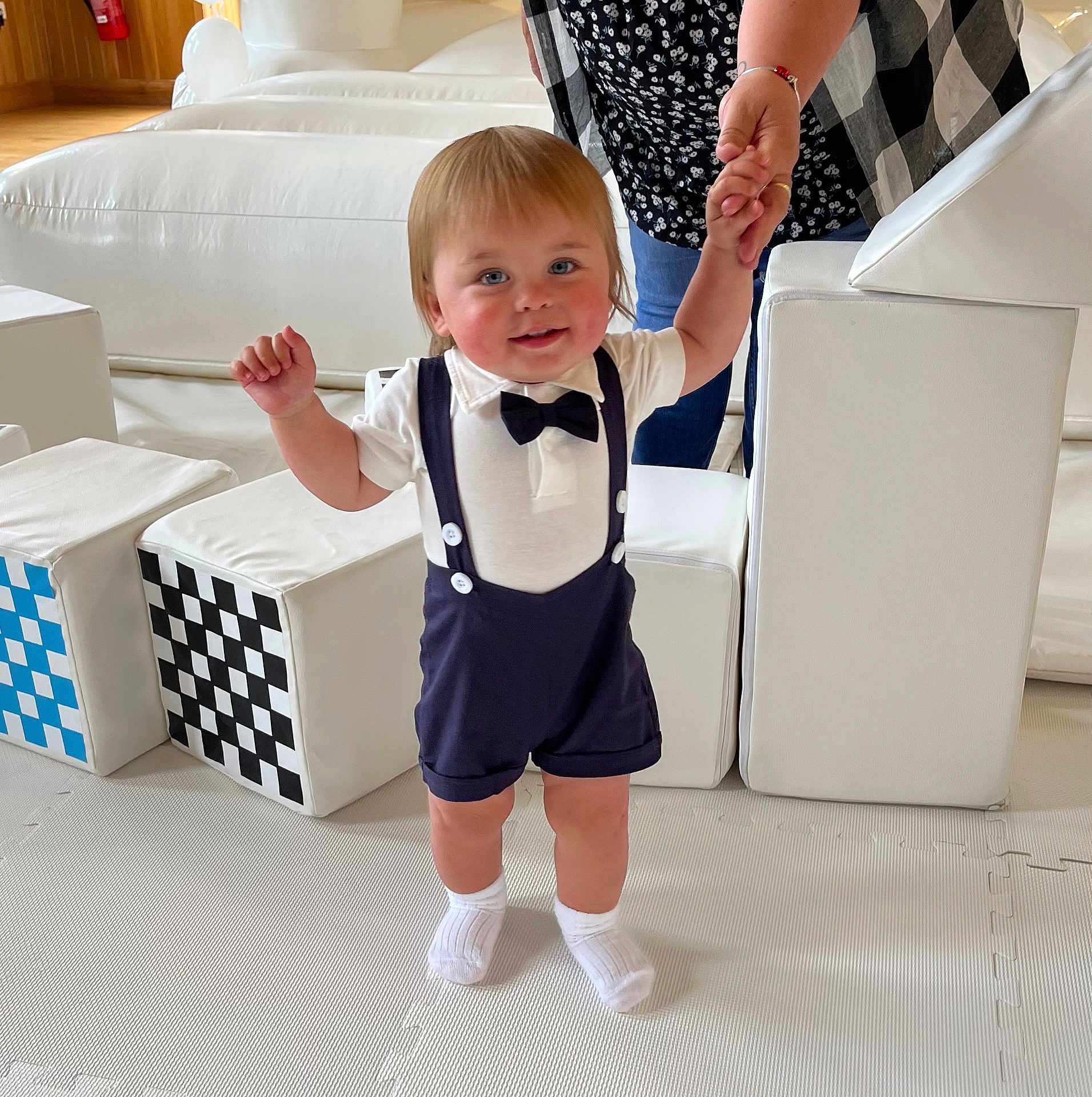 Beau is registered to the contest to win money with this photo: toddler, child, baby, indoor, play_area, white_socks, bow_tie, suspenders, smiling, holding_hand, adult, soft_blocks, checkered_pattern, floor_mat, shorts, cute, walking, person, happy, light