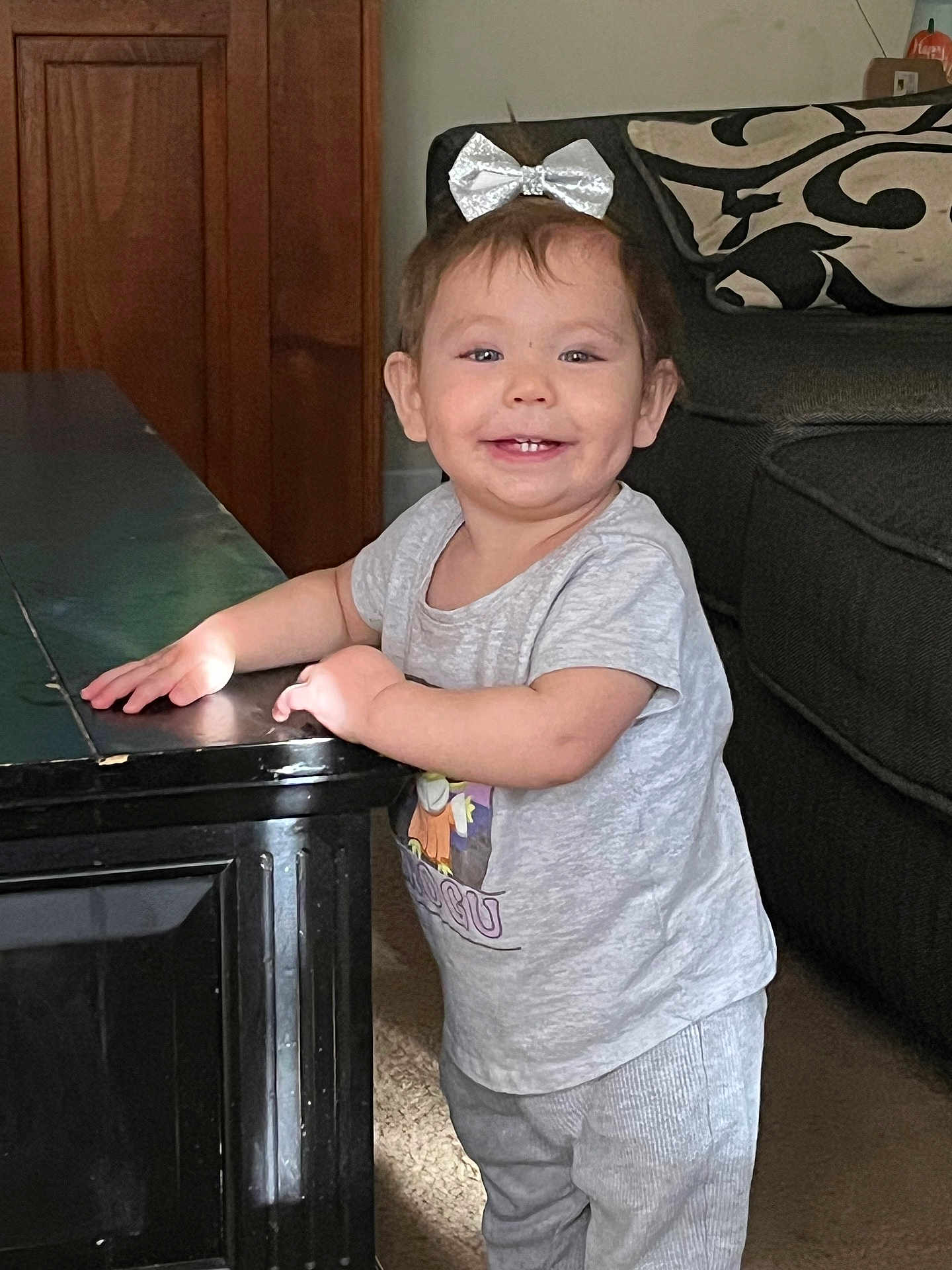 Echõ joined the competition — help win amazing prizes! toddler, child, smile, bow, silver_bow, standing, table, black_table, living_room, couch, patterned_cushion, carpet, short_sleeve_shirt, pants, happy, person, indoor, furniture, cute, baby