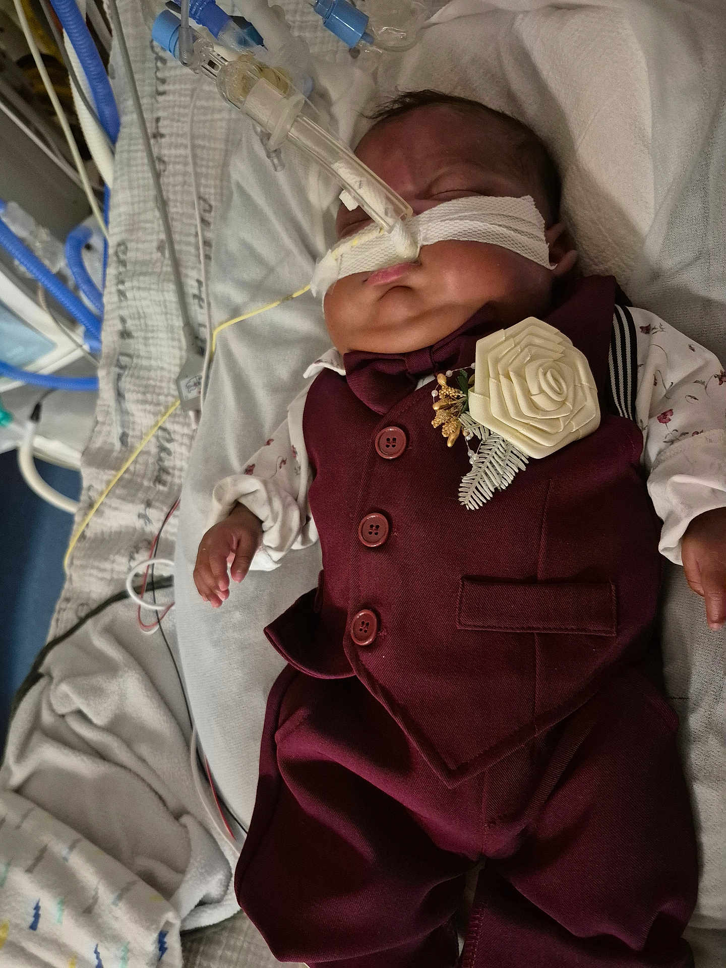 Mateo Marcelin is registered to the contest to win money with this photo: baby, infant, hospital, medical_tubes, nasogastric_tube, maroon_suit, boutonniere, sleeping, bed, blanket, medical_equipment, newborn, child, patient, clothing, face, portrait, healthcare, resting, indoors