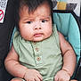 baby, child, infant, green_outfit, sleeveless, wooden_buttons, baby_seat, patterned_background, chubby_cheeks, curly_hair, serious_expression, barefoot, hands, legs, indoor, portrait, close_up, seatbelt, car_seat, person