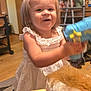 Aspen is registered to the contest to win money with this photo: toddler, child, white_dress, toy_gun, rocking_horse, indoor, smiling, happy, blond_hair, furniture, carpet, wood_floor, vase, flowers, walker, home, person, cute, playful, young_child