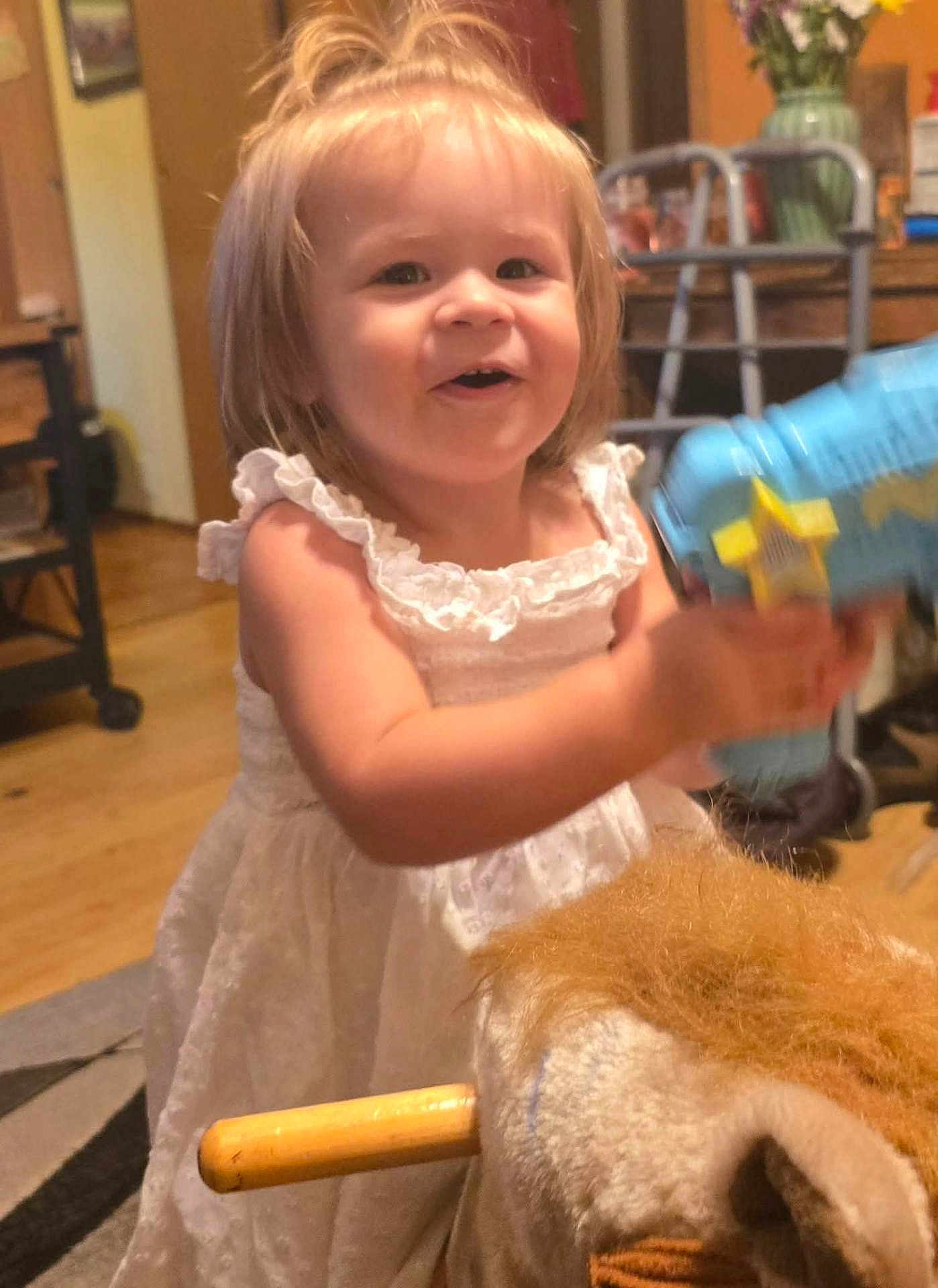 Aspen is registered to the contest to win money with this photo: toddler, child, white_dress, toy_gun, rocking_horse, indoor, smiling, happy, blond_hair, furniture, carpet, wood_floor, vase, flowers, walker, home, person, cute, playful, young_child