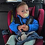 toddler, child, car_seat, pacifier, blue_jacket, toy, blanket, car_interior, seatbelt, footwear, window, headrest, clothing, person, young_child, casual, indoor, safety, travel, cute