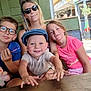 child, children, woman, glasses, cap, pink_clothing, blue_clothing, plush_toy, table, outdoor, cafe, smile, happy, person, sunlight, sunglasses, bench, tattoo, street, barrel