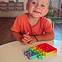 blue_eyes, bracelet, casual, child, furniture, happy, indoor, natural_light, orange_shirt, person, playroom, pop_it, rainbow, short_hair, smiling, table, toddler, toy, window, wooden_floor